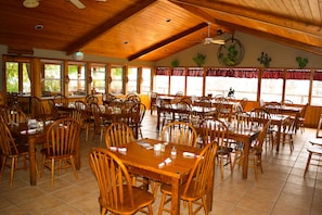 A spacious dining area with wooden tables and chairs, large windows, and a high ceiling with wooden beams.