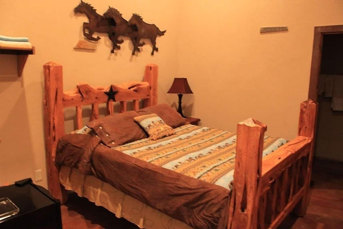 Una cama de madera con cabecera, una colcha estampada y almohadas. Hay una lámpara y un adorno de caballos en la pared.