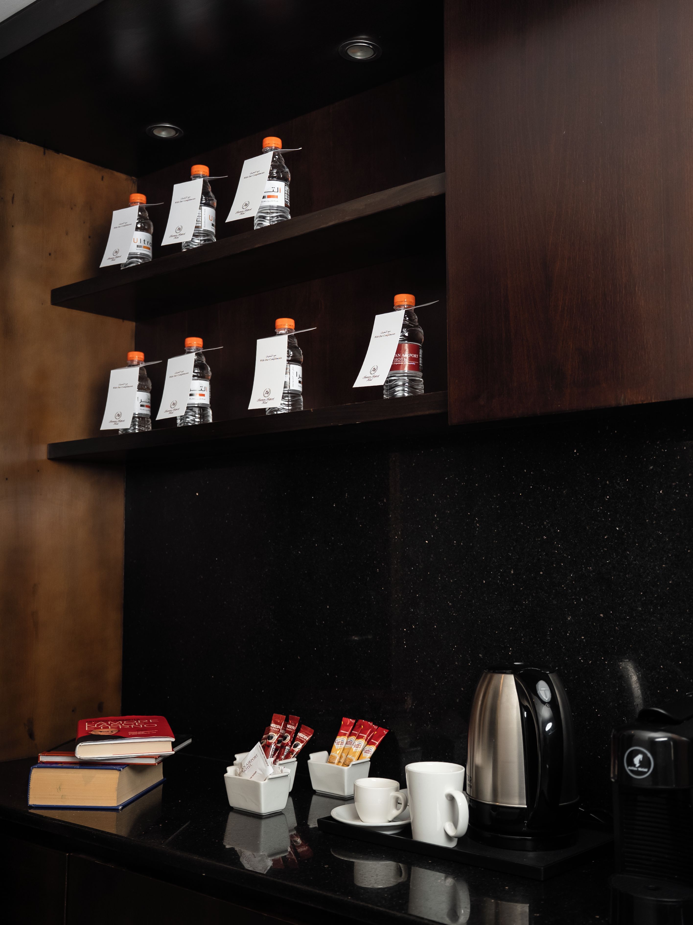 Habitación de hotel con una encimera negra, una cafetera, una taza, un libro y estantes con botellas de agua.