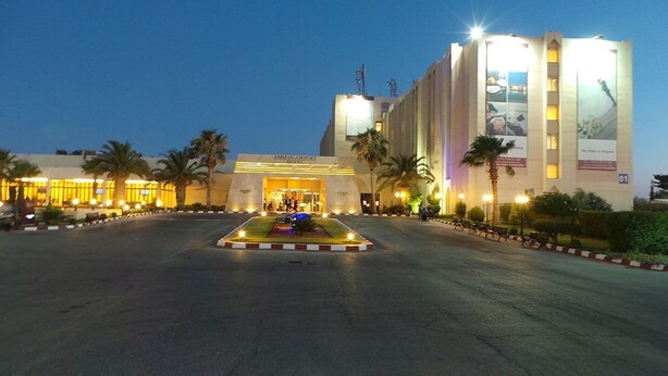 Entrada de hotel con fachada iluminada, palmeras y un jardín central.