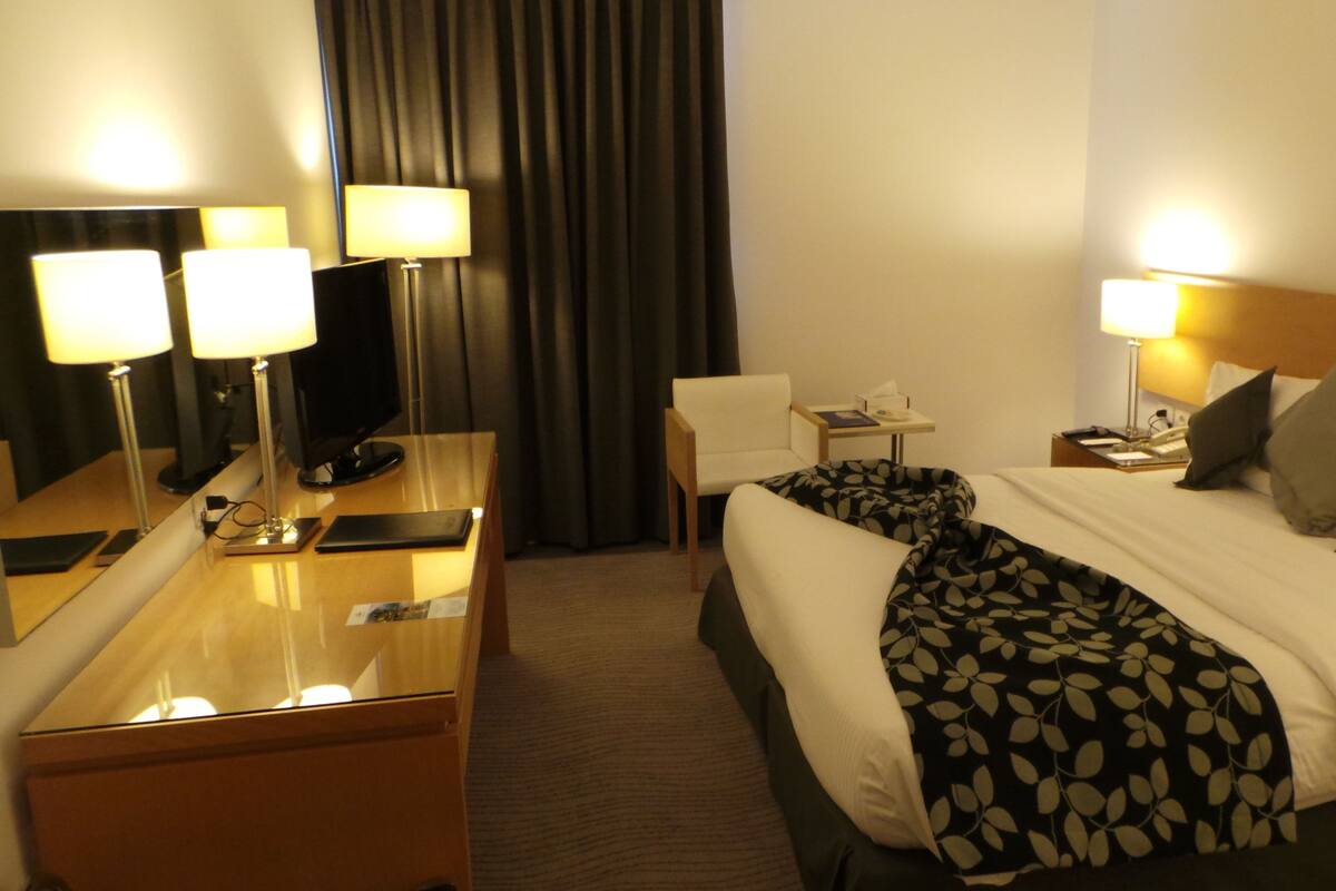 A hotel room with a bed, bedside table, lamp, chair, and a desk with a computer and phone.