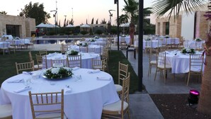 Un espacio formal para comer al aire libre con mesas redondas, manteles blancos y sillas de madera, con vista a una piscina y palmeras.