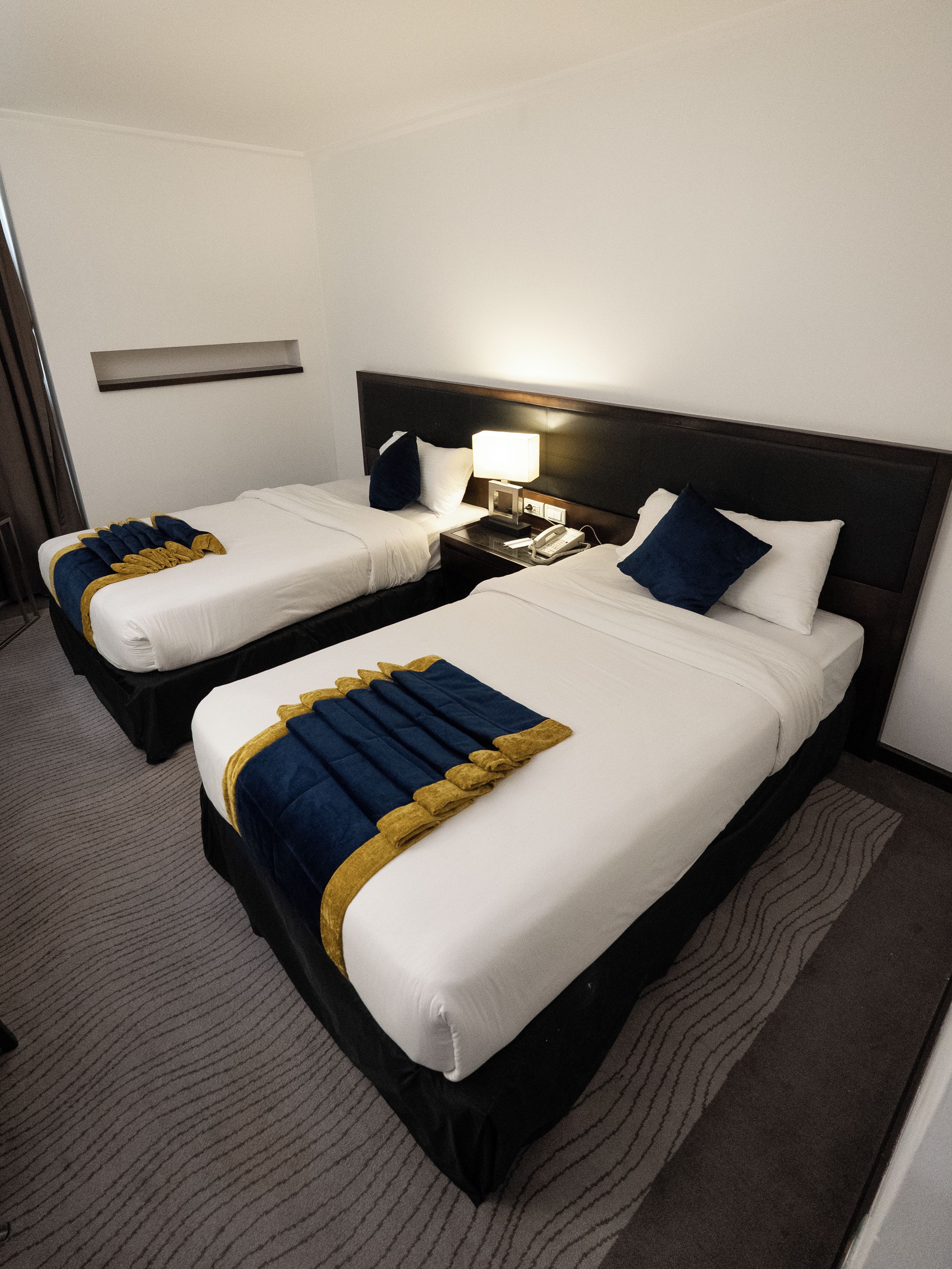 Quarto de hotel com duas camas, mesa de cabeceira com abajur e prateleira fixada na parede.
