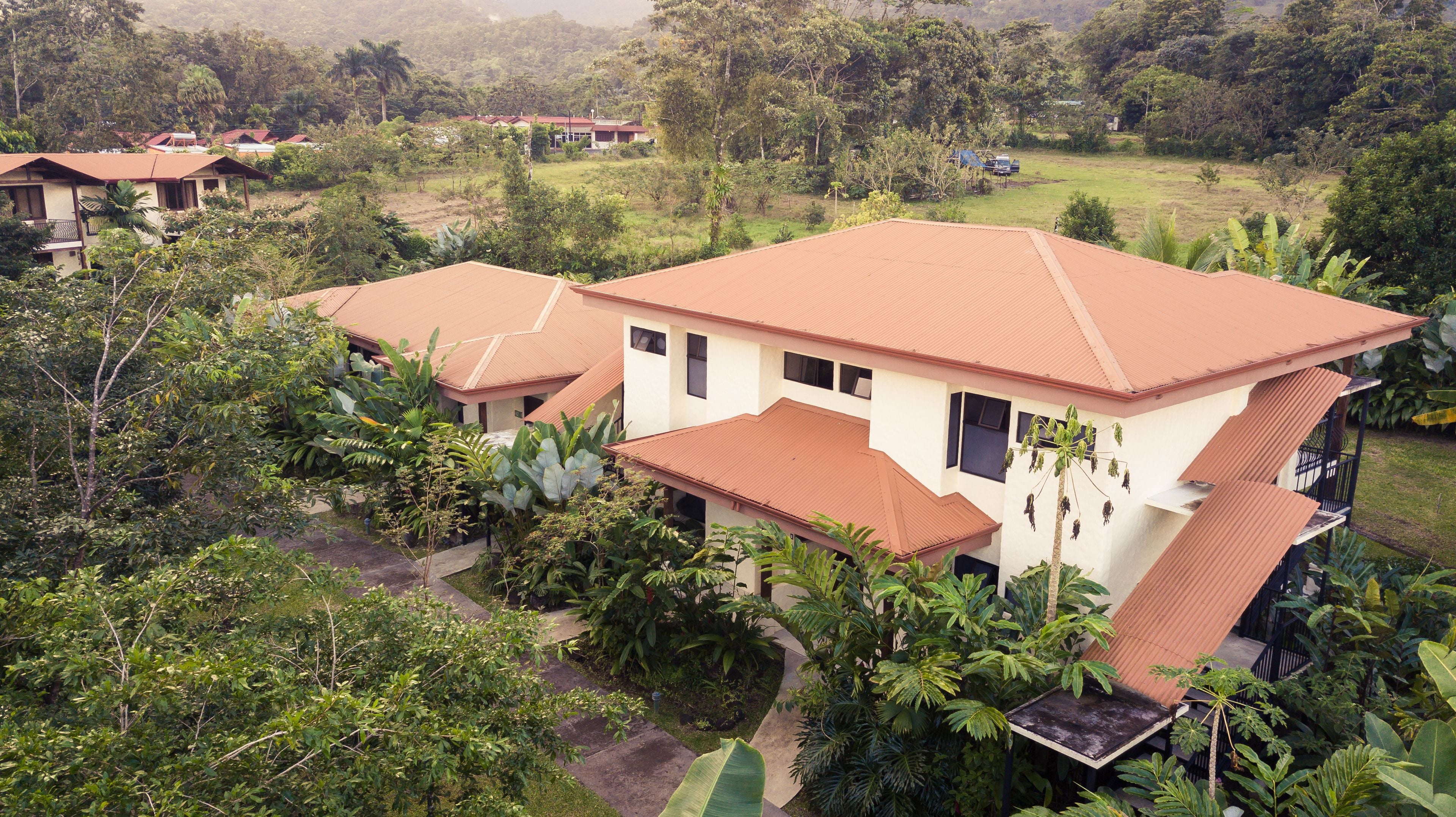 A two-story house with a red roof, surrounded by lush greenery and other houses in the background.
