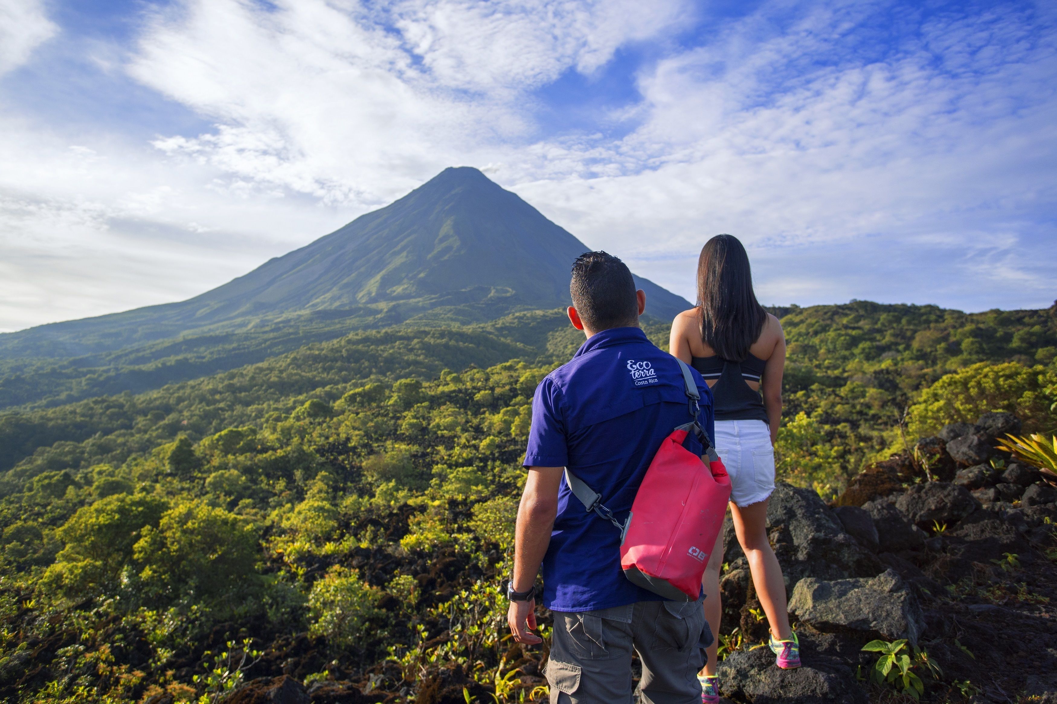 Two people standing on a rocky outcrop overlooking a lush, green landscape with a mountain in the background.