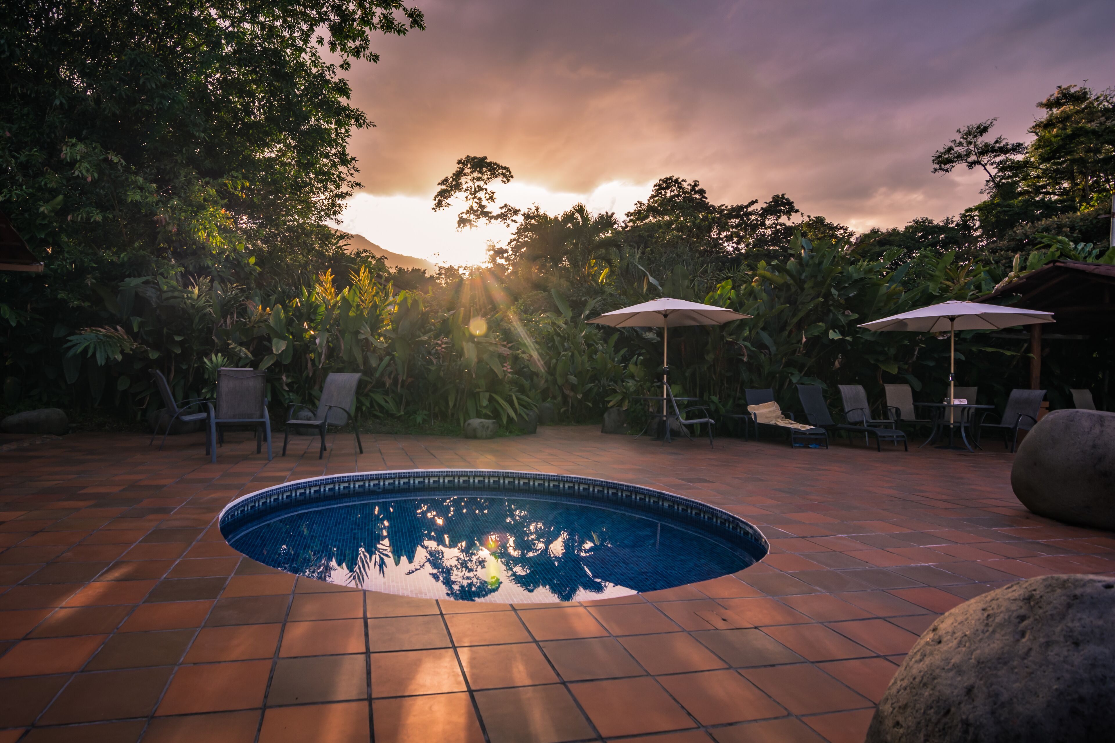 A swimming pool with tiled flooring, surrounded by chairs and umbrellas, set against a backdrop of trees and a sunset.
