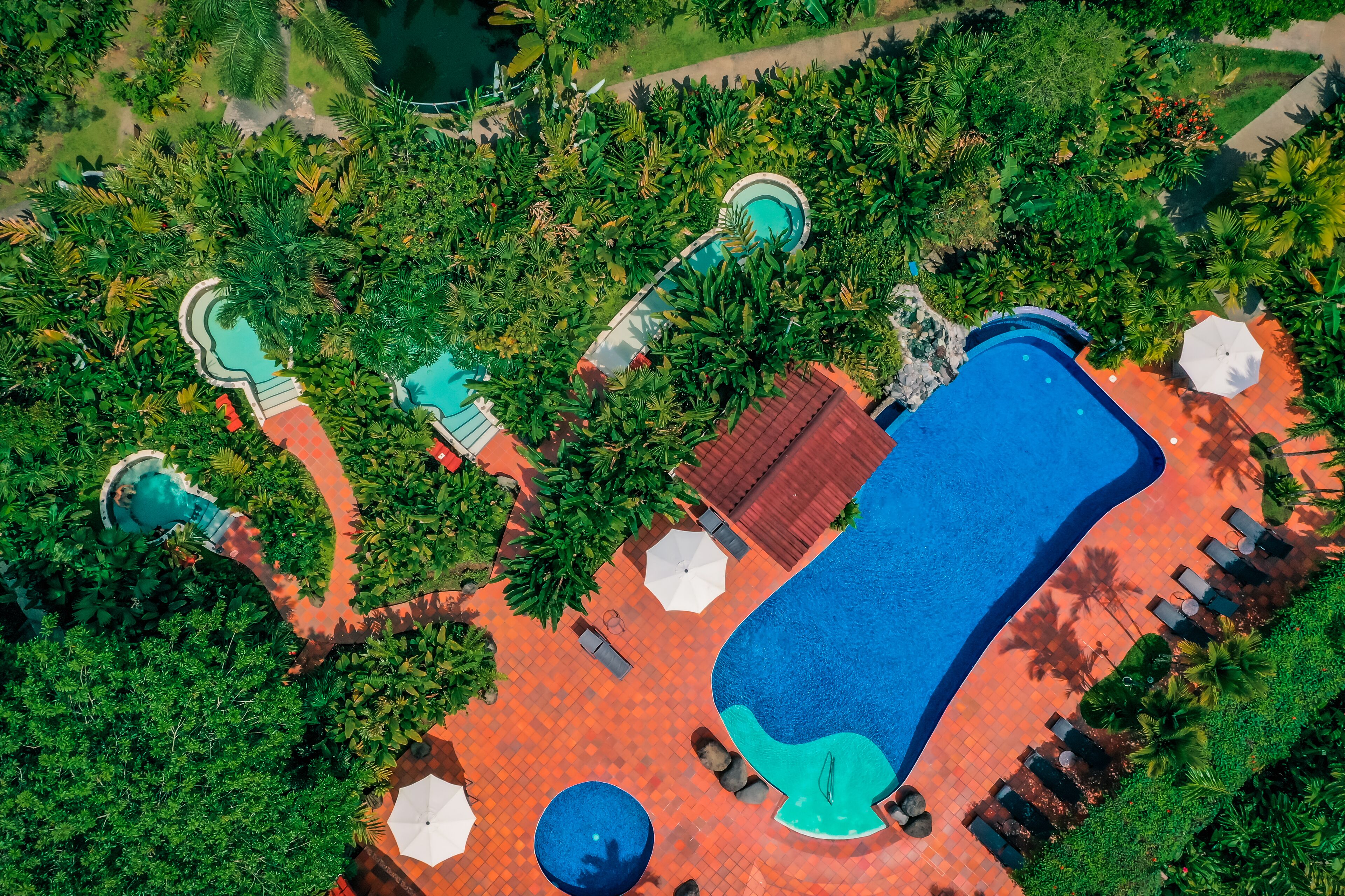 An aerial view of a resort with multiple swimming pools, surrounded by lush greenery and palm trees.