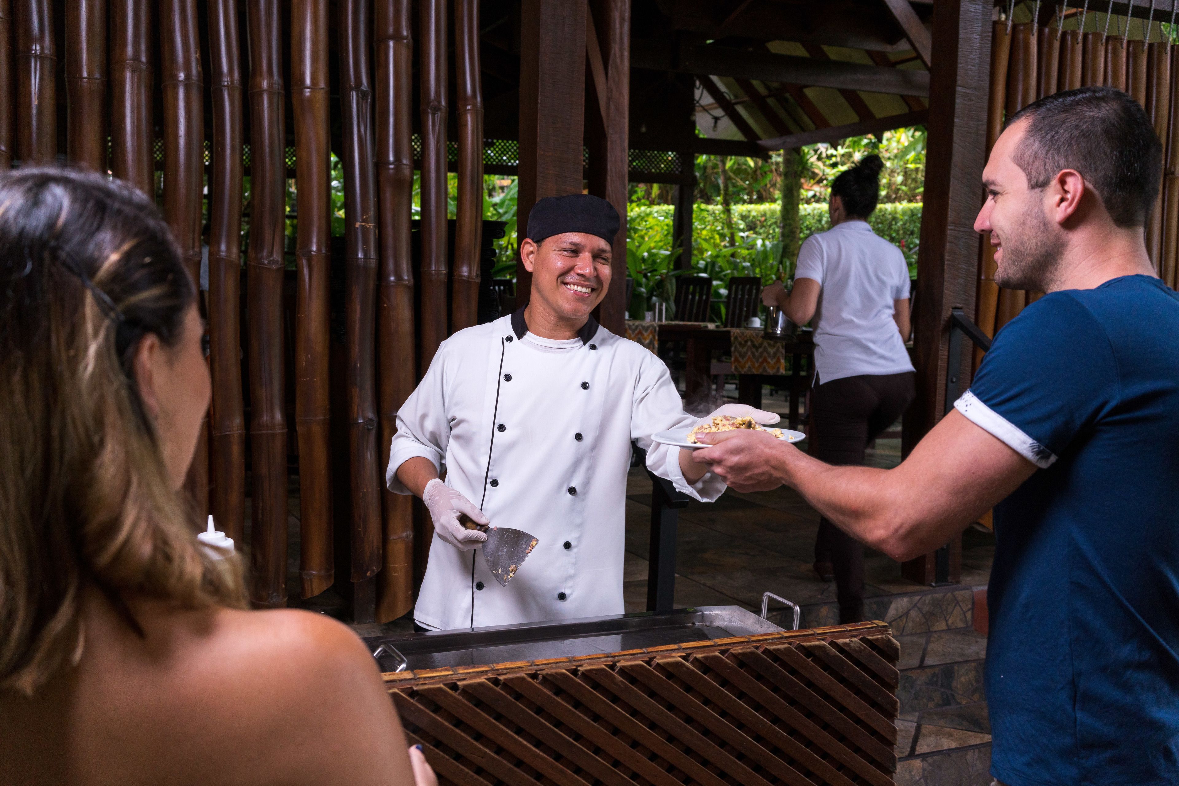 A chef serving food to a customer at an outdoor restaurant with bamboo walls and a covered seating area.