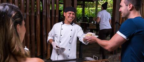 A chef serving food to a customer at an outdoor restaurant with bamboo walls and a covered seating area.
