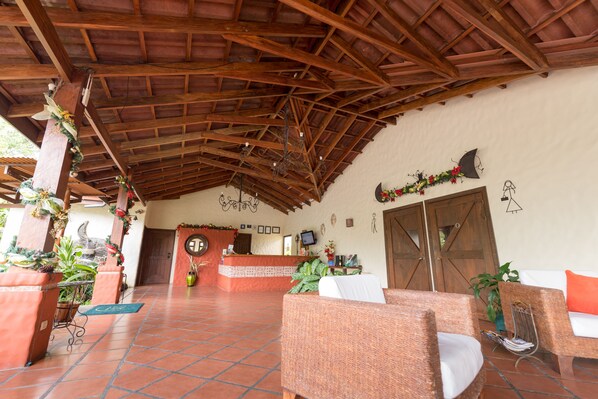 A reception area with a tiled floor, wooden ceiling beams, wicker furniture, and a reception desk with a computer monitor. - Casa Luna Hotel & Spa (La Fortuna)