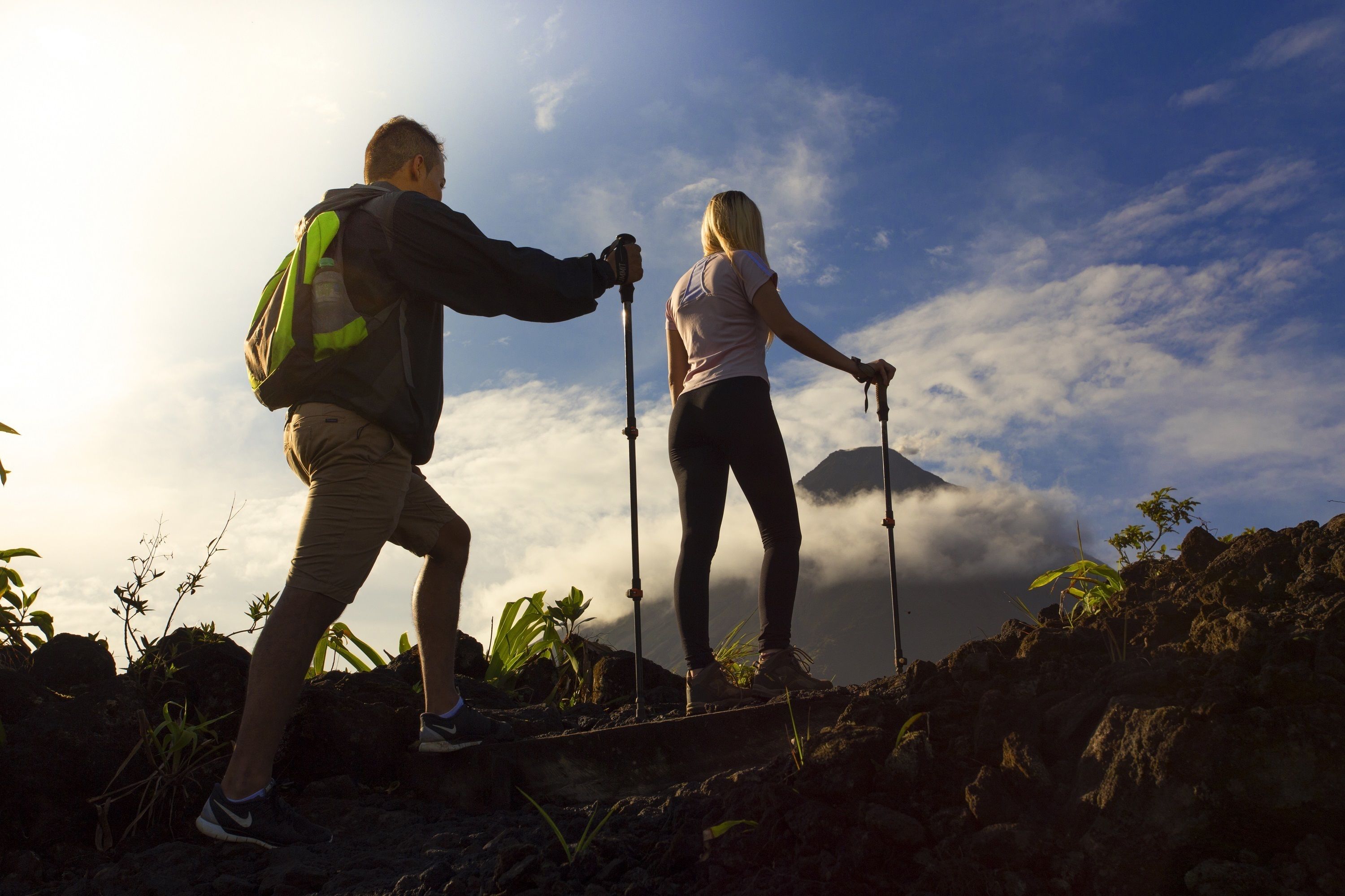 Two people hiking on a rocky path with a mountainous backdrop and a partly cloudy sky.