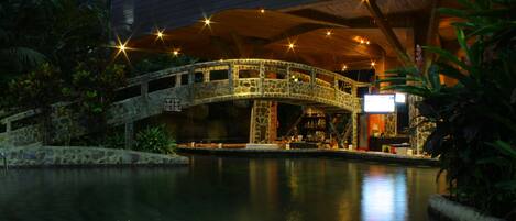 A stone bridge over a reflective pool, leading to a building with a thatched roof and illuminated interior.