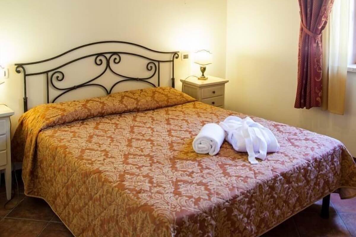 A bedroom with a bed, a nightstand, a lamp, a chandelier, and a window with curtains.