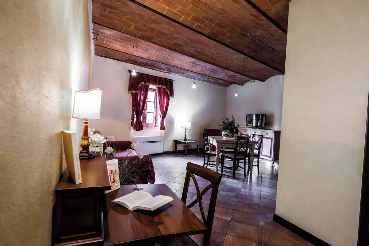 A cozy room with a wooden table, a chair, a lamp, and a book. There is a window with curtains and a view of a kitchen area.