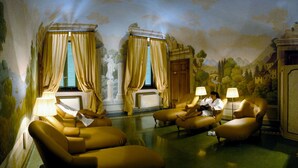 A room with two yellow armchairs, a sofa, and a table with lamps. There are two windows with curtains, and a mural on the wall.