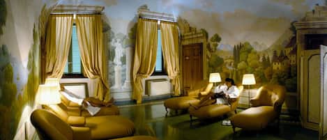 A room with two yellow armchairs, a sofa, and a table with lamps. There are two windows with curtains, and a mural on the wall.