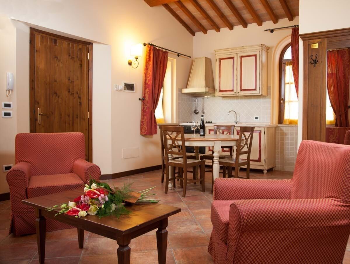 Una sala de estar acogedora con sillones tapizados en rojo, una mesa de centro de madera con un arreglo floral, y una cocina al fondo con armarios de madera y salpicadero de azulejos.