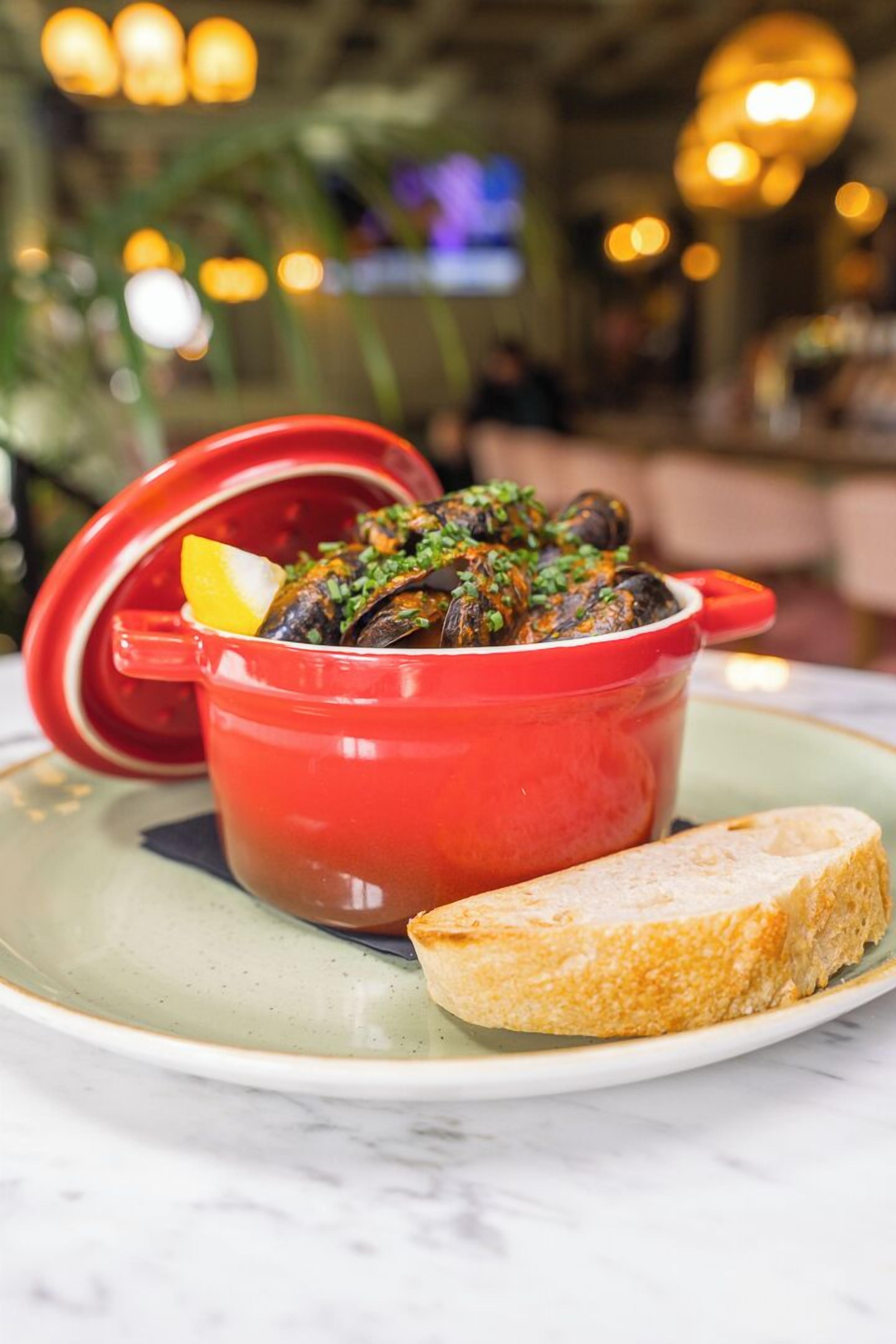 A red pot of mussels with a lemon wedge on a plate, accompanied by a piece of bread.