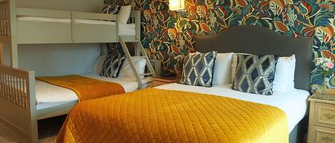 Una habitación con dos camas, una literas y otra individual, ambas con colchas. Hay una mesita de noche con lámpara, un armario y un papel tapiz con estampado floral.
