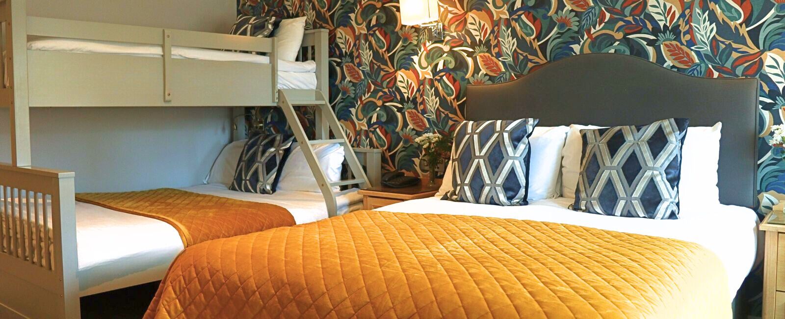 Una habitación con dos camas, incluyendo una litera, un papel tapiz floral y una mesita de noche con una lámpara.