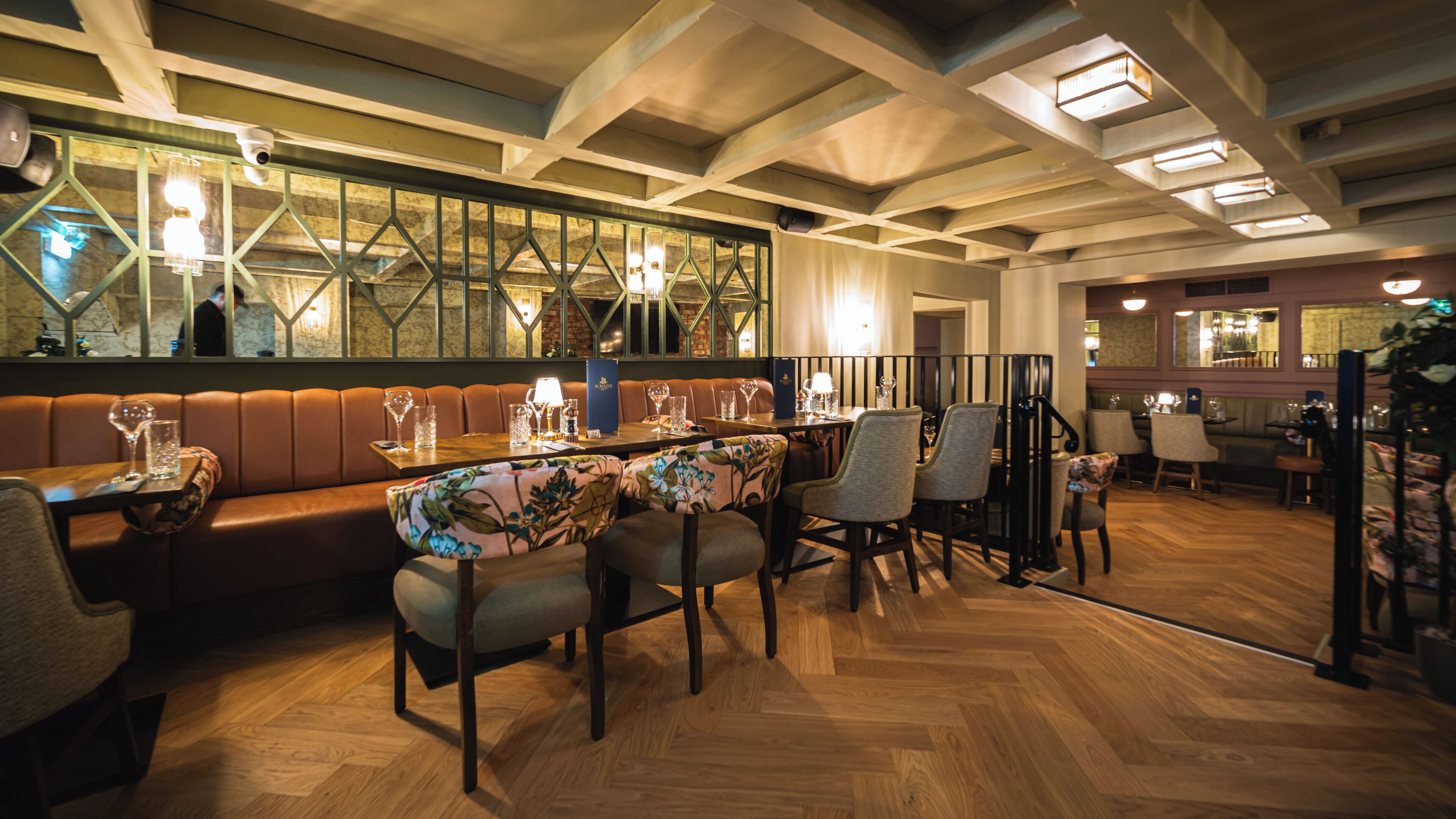 A restaurant interior with a wooden herringbone floor, leather booth seating, and a bar area with a high ceiling and exposed beams.