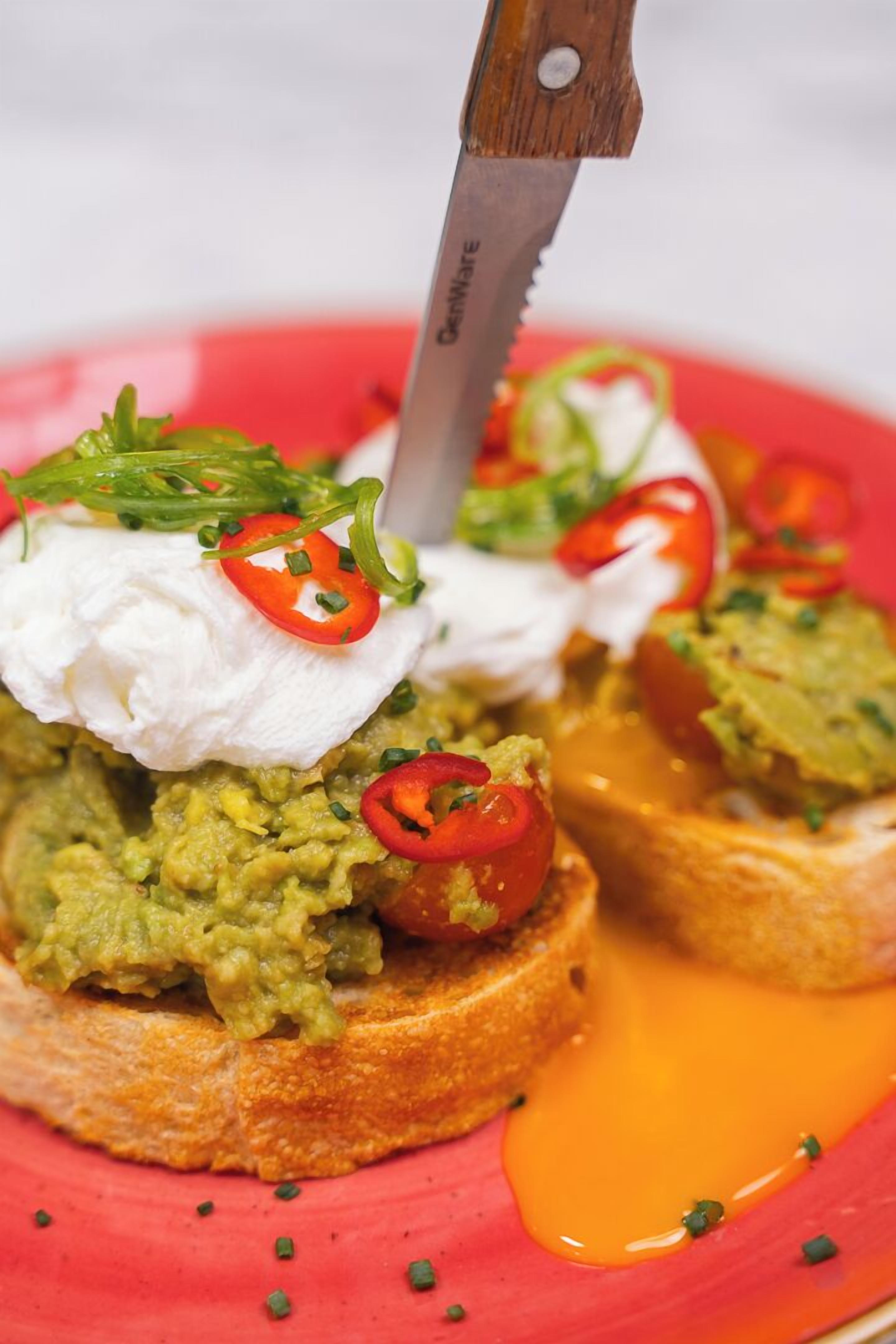 Un plato con tostada de aguacate cubierto con crema agria, rodajas de chile rojo y cebollines.