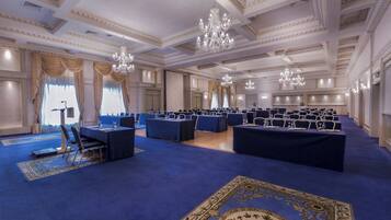 Une salle de conférence officielle, avec des rangées de tables et de chaises, des lustres et des détails décoratifs élaborés au plafond.