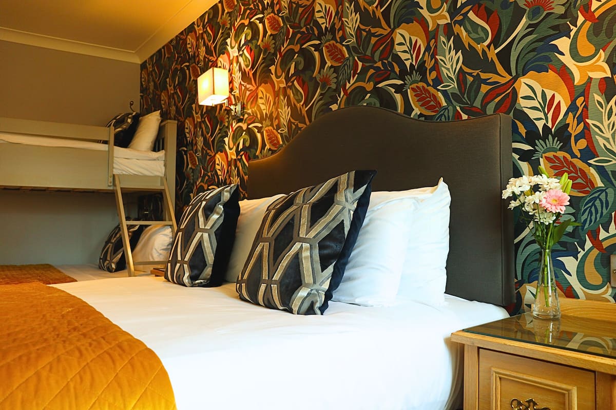 Une chambre d’hôtel avec un lit superposé, un grand lit agrémenté d’oreillers à motifs, une table de chevet avec un vase de fleurs et un mur recouvert d’un papier peint à motifs floraux.