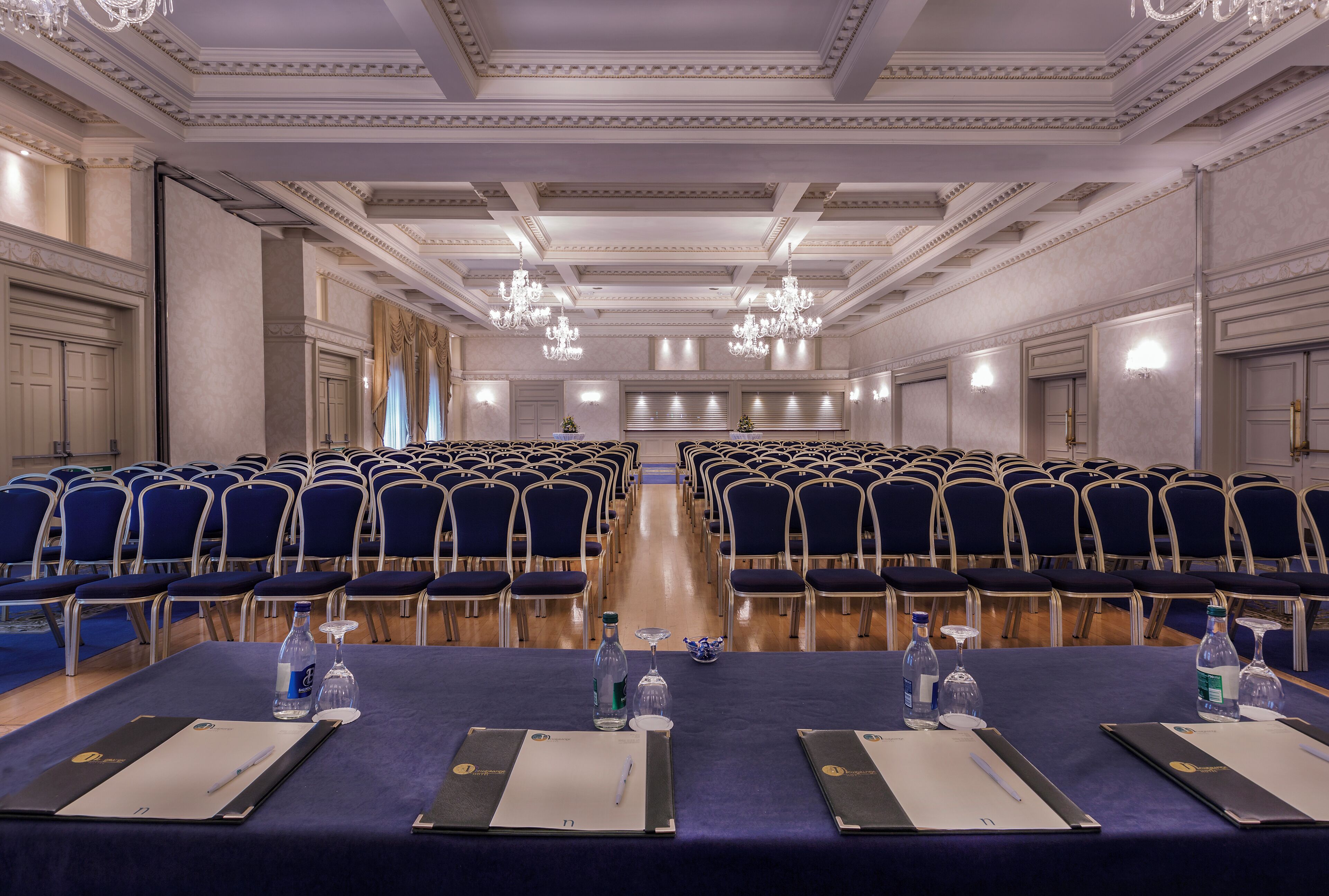 A conference room with rows of chairs, tables with water bottles, and a chandelier.