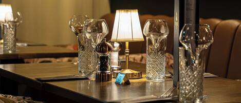 A dining table set with crystal glassware, a lamp, and a menu stand in a warmly lit room with a geometric wall design.