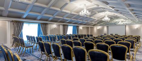 Un salón de conferencias con filas de sillas, una araña de cristal y una alfombra estampada.
