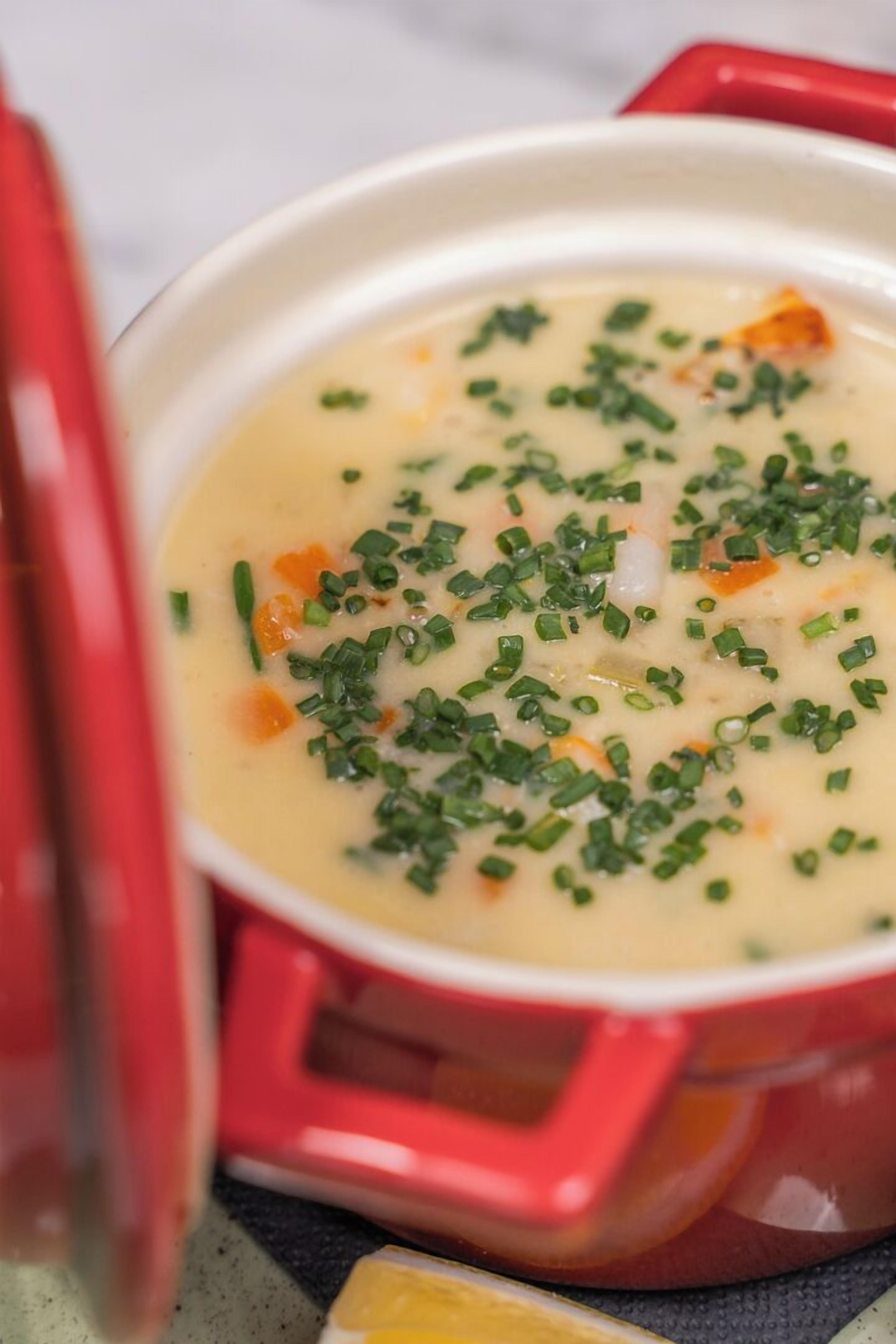 Un pot rouge contenant une soupe crémeuse, garnie d’herbes vertes hachées.
