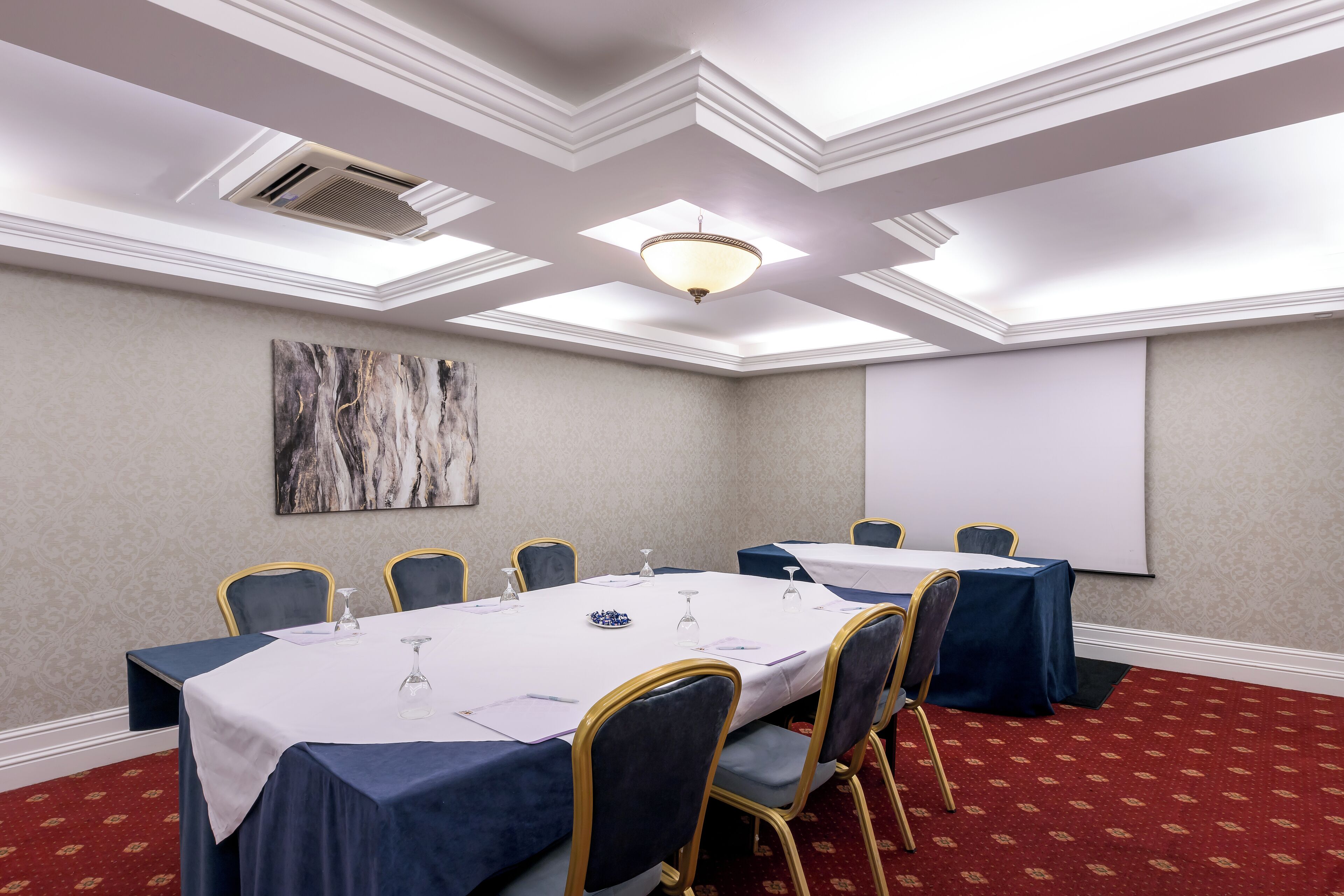 Un salón de conferencias con una mesa blanca, sillas azules y una pantalla de proyector en la pared.