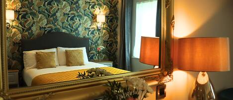 A hotel room with a bed, a mirror, a lamp, and a floral arrangement.