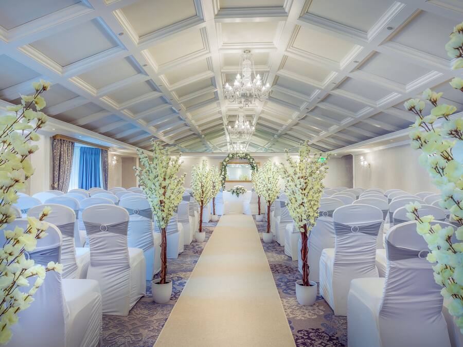 Un espacio para eventos formales con hileras de sillas blancas, un pasillo central y flores blancas decorativas.