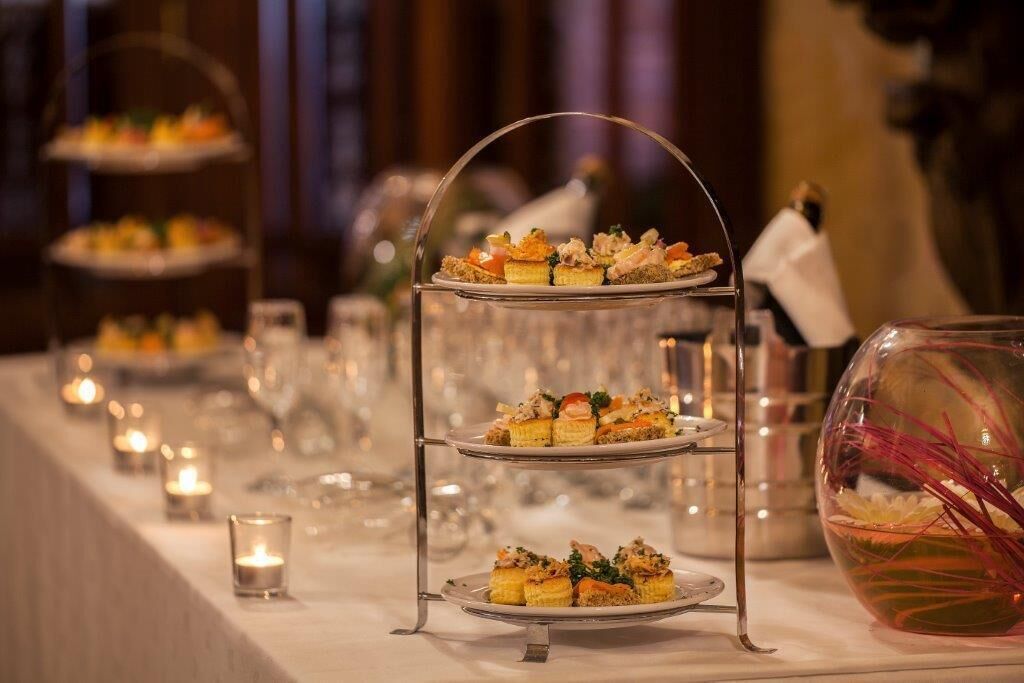 A tiered dessert stand with multiple layers of pastries, a glass vase with red flowers, and candles on a table.