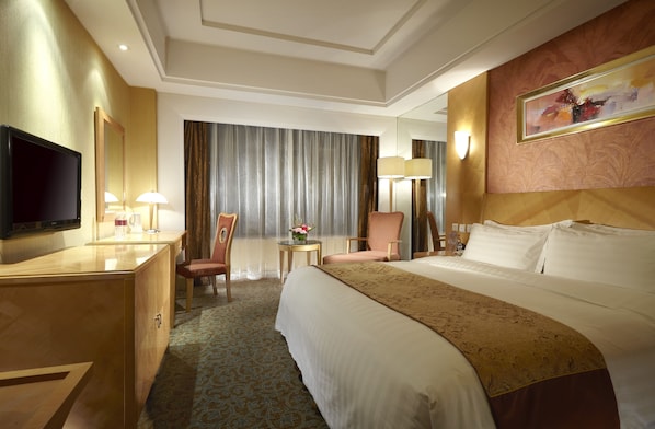 Habitación de hotel con una cama grande, un escritorio con televisor, una silla, una mesita con un jarrón de flores y un ventanal con cortinas.
