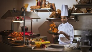 Un chef dans une cuisine professionnelle, entouré de divers ustensiles de cuisine, d’une planche à découper et d’une variété de produits de boulangerie disposés sur des étagères à l’arrière-plan.