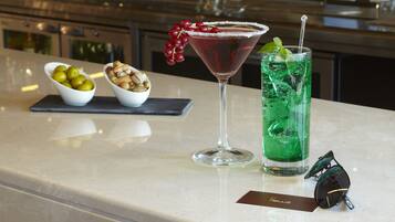 A bar counter with a martini glass and a tall glass with a straw, two bowls of olives and nuts, and a bottle of liquor in the background.