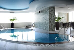 Ein überdachter Schwimmbereich mit klarem Wasser, umgeben von Fliesenboden und einer Decke mit Sternenmuster. In der Nähe des Pools befinden sich Stühle und ein Tisch, und rundherum sind Blumentöpfe aufgestellt.