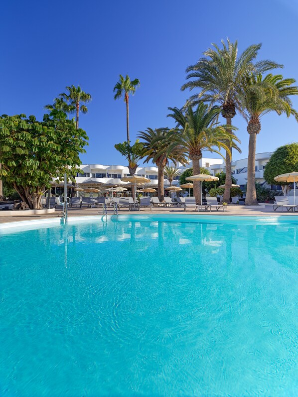 A clear blue pool with lounge chairs and umbrellas, surrounded by palm trees and a building with a white facade. - H10 Ocean Dunas - Adults Only (La Oliva)