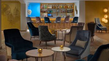 Un área de lounge moderna con diversas opciones de asientos, un bar con amplia selección de licores y una pared decorativa con arte abstracto.