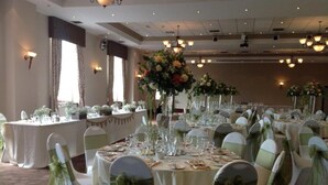 A banquet hall with round tables, white chairs with green sashes, and tall floral centerpieces. - The Towers Hotel and Spa (Neath)