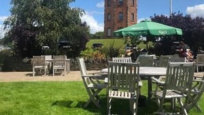 An outdoor seating area with tables and chairs, a green umbrella, and a brick tower in the background. - The Towers Hotel and Spa (Neath)