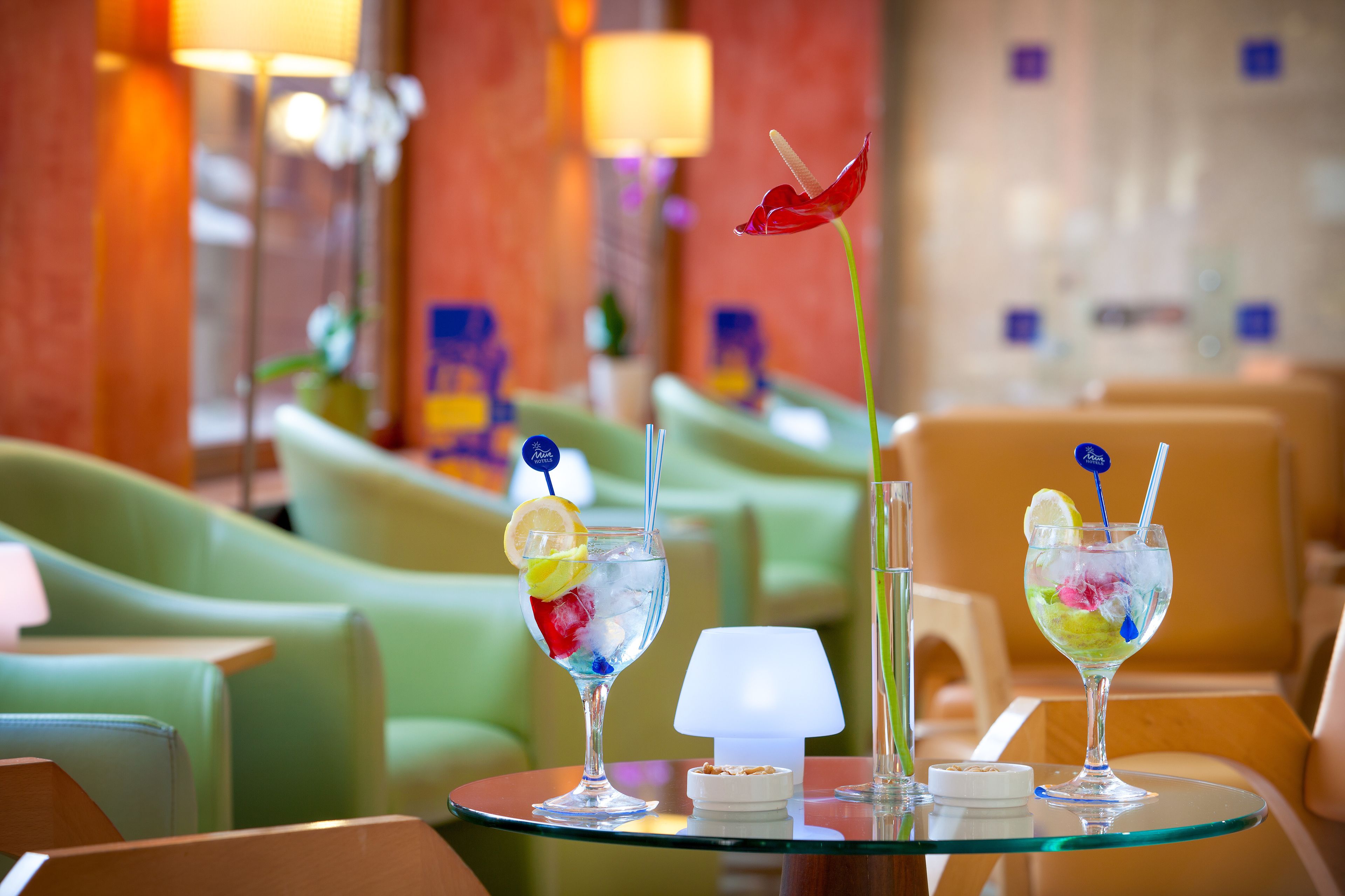 Two cocktails on a glass table with a lamp and a small bowl, in a room with green chairs and a red wall.