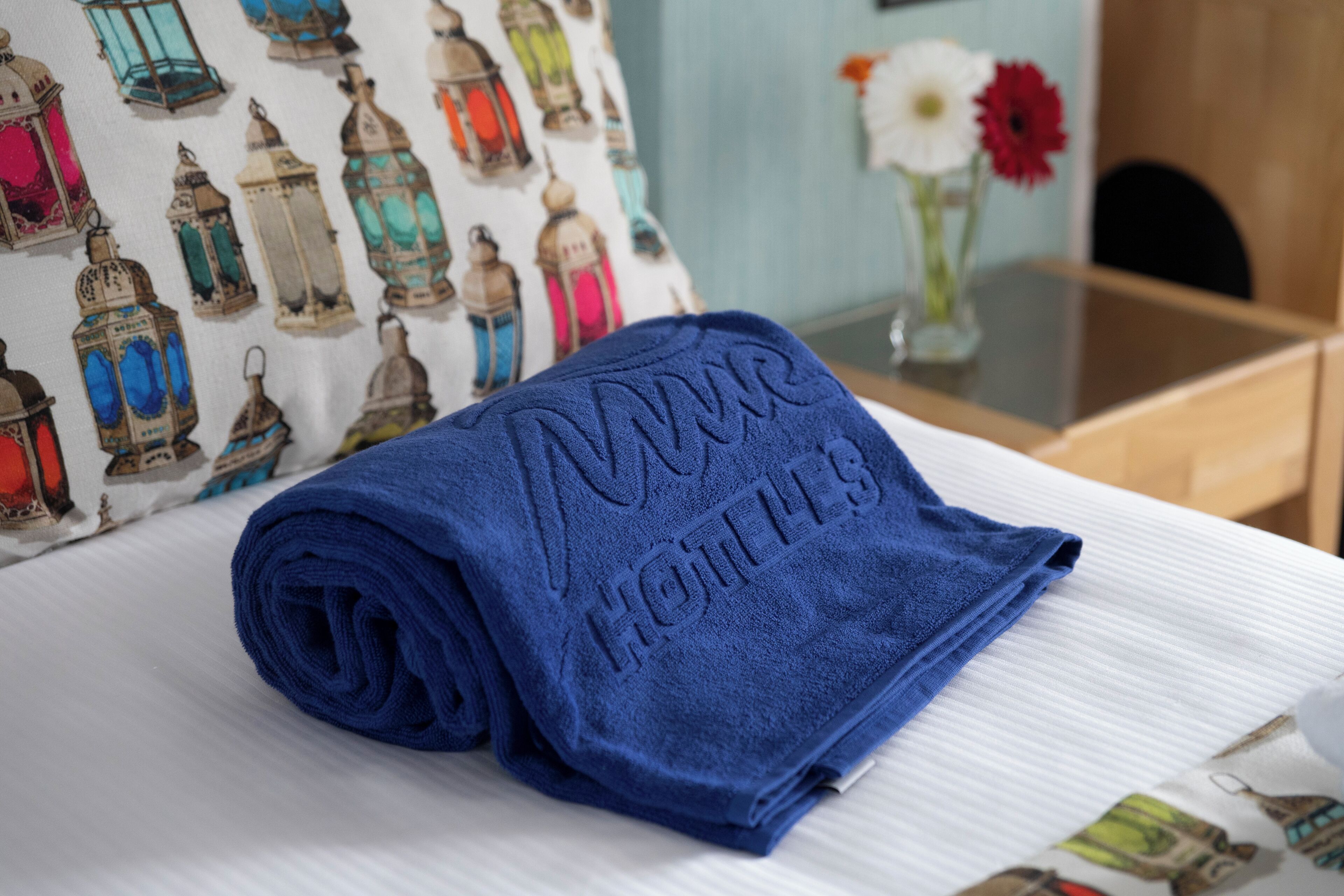A neatly folded blue towel with "MOKO HOTELS" embossed on it, placed on a bed with a white sheet and a pillow.