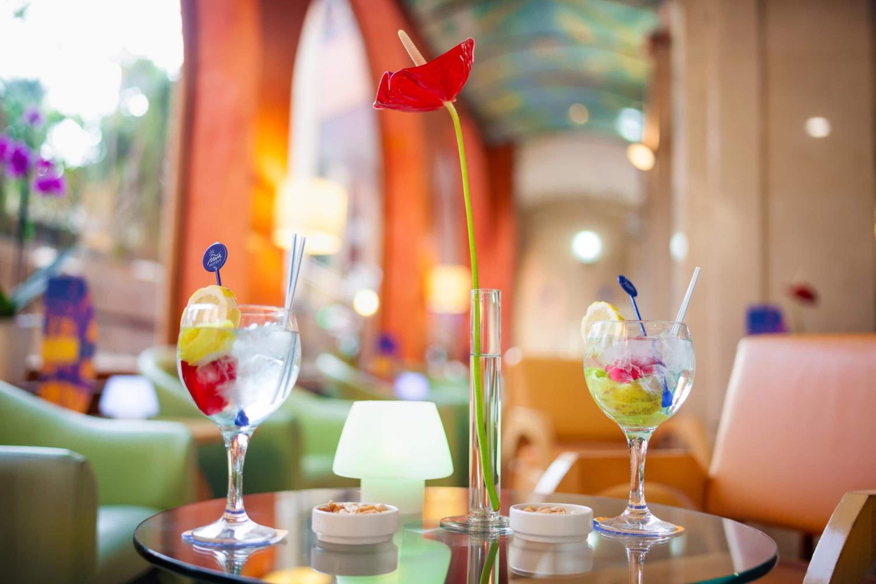Three cocktails on a table with a lamp and small bowls, in a brightly lit indoor setting.