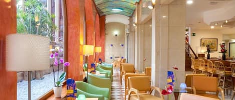 Un vestíbulo de hotel con piso a cuadros, asientos en tonos verde y beige, una mesa de vidrio con bebidas y un mural de techo colorido.