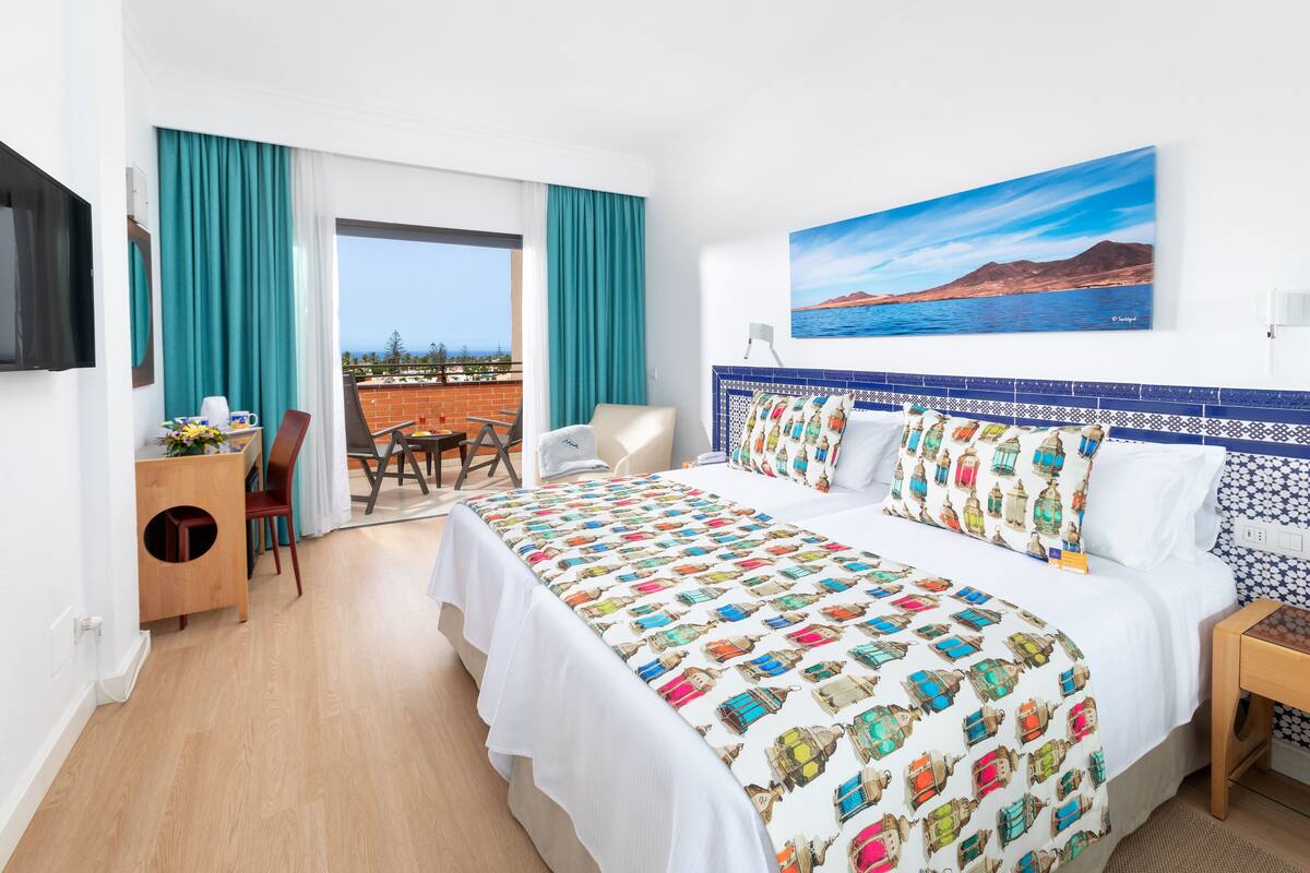 Une chambre d’hôtel avec un grand lit, un bureau avec une chaise, une télévision et un balcon donnant sur la mer.