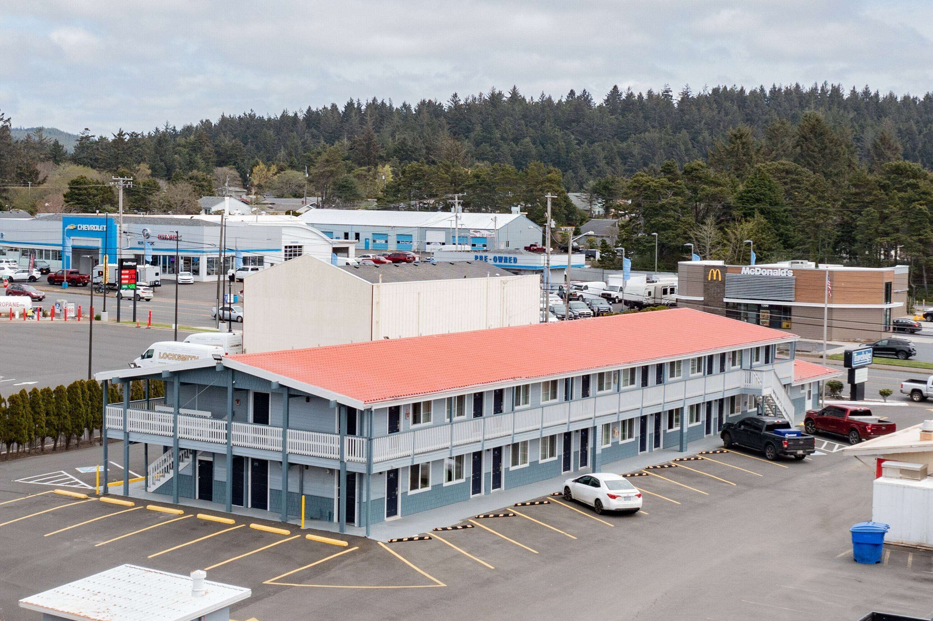A motel with a red roof, multiple parking spaces, and a view of a forested hill in the background.